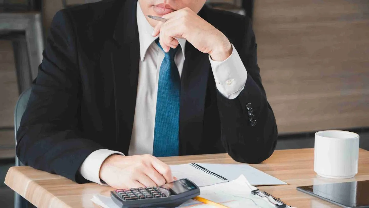 A man in a black suit is stationed at his desk with a calculator and papers