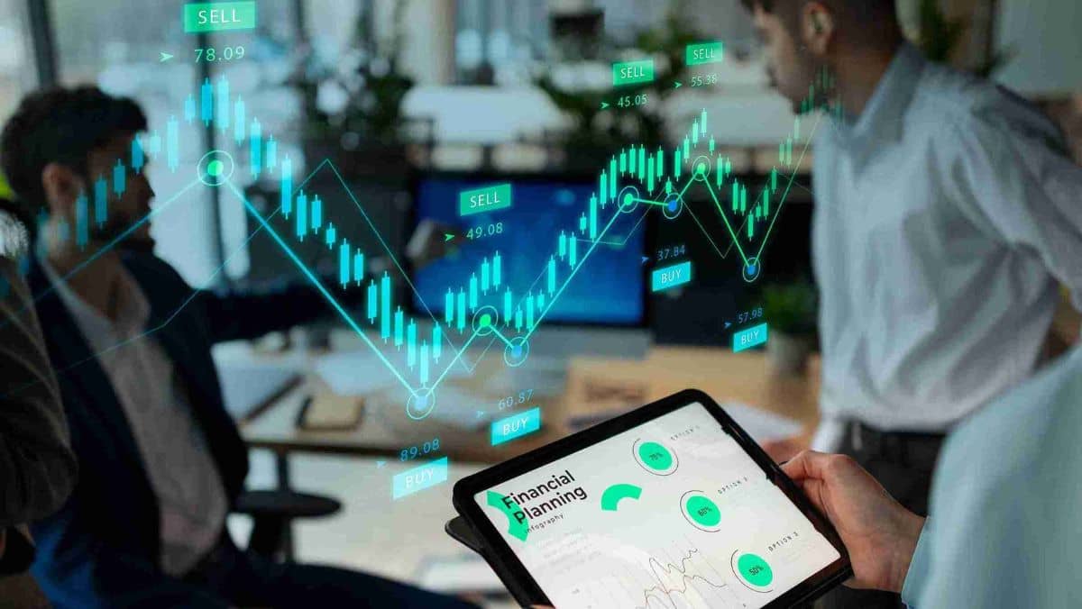 A man in the background holds a tablet surrounded by others, while a graphical representation of a trading chart is displayed