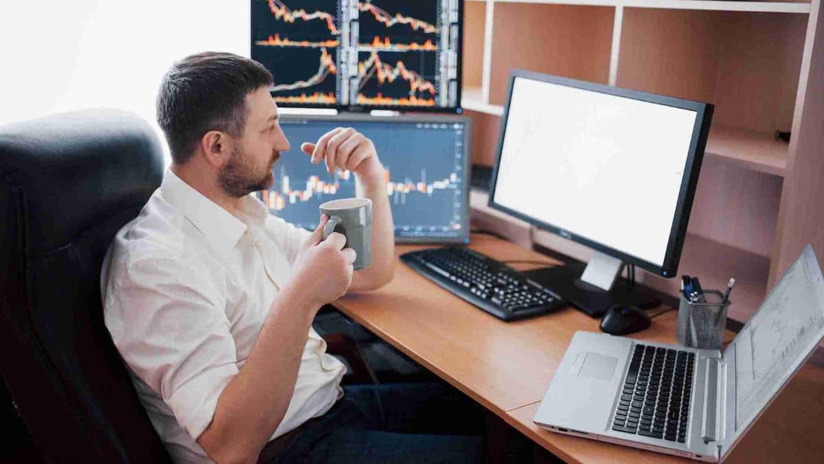 A man with a cup of coffee is looking at multiple computer screens