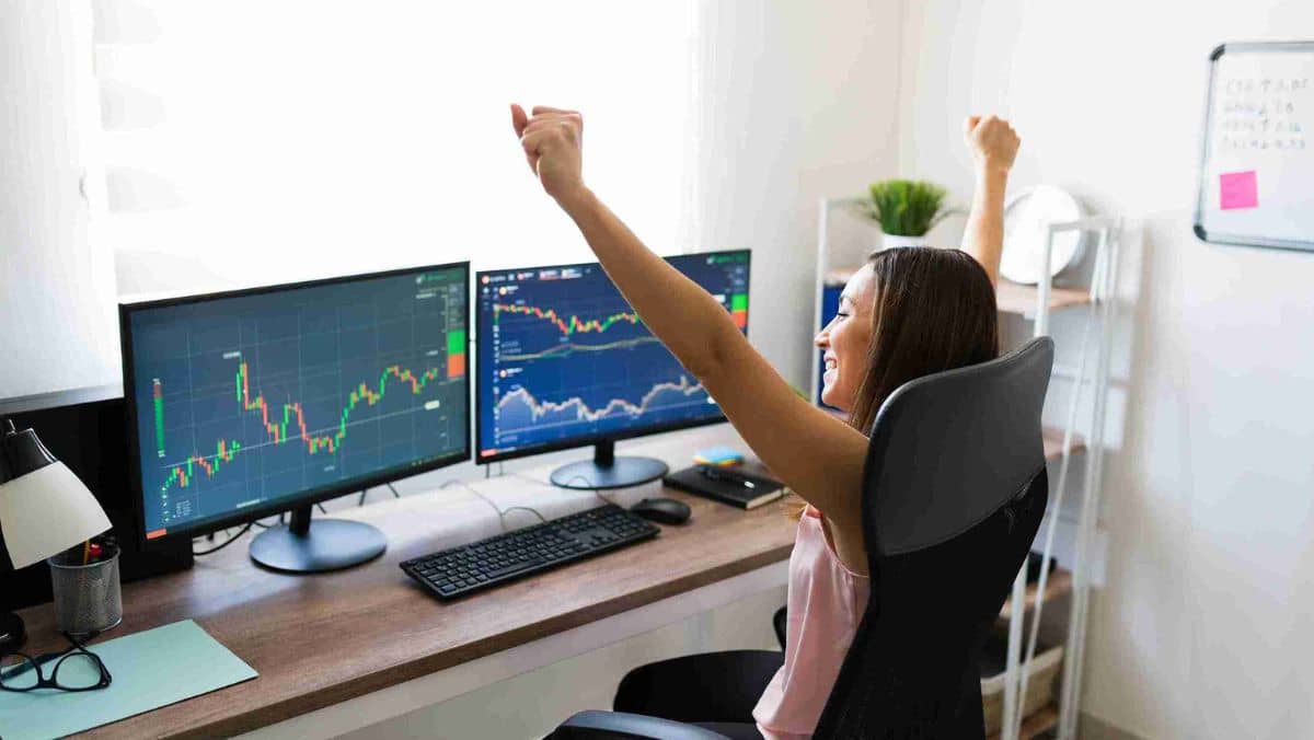 A woman celebrating with her hands raised while looking at multiple computer monitors