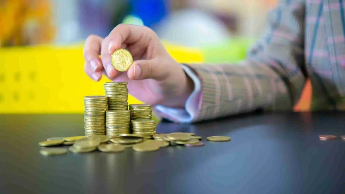 Hand holding a gold coin over a stack of gold coins on a table